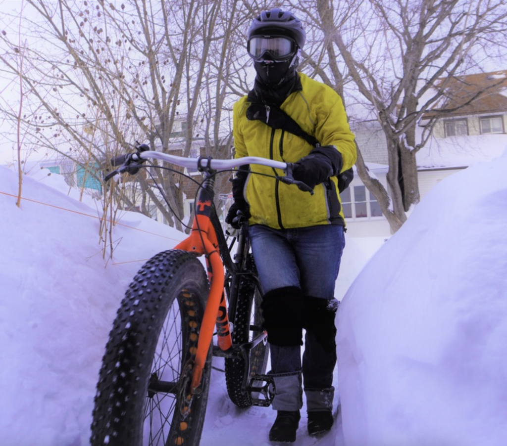 Person in yellow jacket, ski goggles and helmet with orange fat bike wheeling down snow lined path